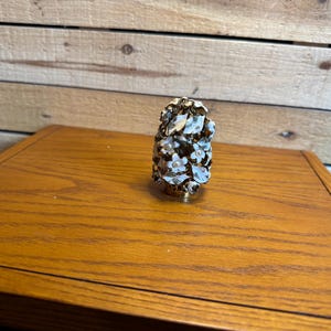 Puede incluir: Un tapón decorativo para botellas con una base de metal dorado y un grupo de formas florales blancas con pequeños centros perlados y hojas metálicas. El arreglo floral es denso y texturizado, creando una forma escultórica redondeada.