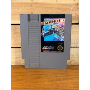 以下が含まれることがあります： Tiger-HeliのNintendo Entertainment System用グレーのビデオゲームカートリッジ。ラベルは黒い背景で、金と白の文字でタイトルが書かれ、黒いヘリコプターのイラストとAcclaimおよびNintendoのロゴが配置されています。