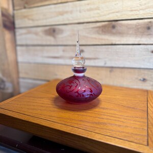 May include: A small, round, deep red glass perfume bottle featuring a frosted finish with an intricate, swirling etched pattern. The bottle is topped with a clear, pointed glass stopper that has a spherical base and a long, slender spire.