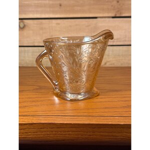 May include: A marigold-colored iridescent glass creamer featuring a raised floral and leaf pattern across its exterior surface. The vessel includes a curved handle on one side and a small pouring spout at the top rim, showcasing a translucent finish.