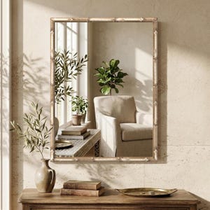 May include: A rectangular wall mirror featuring a light-toned, bamboo-style frame. The reflective surface displays a room interior with a beige armchair, a potted plant, and a window, creating a clear, detailed reflection of the space.
