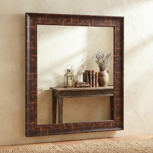 May include: A large rectangular wall mirror with a dark brown frame featuring a distressed, textured finish. The mirror reflects a wooden console table holding a metal lantern, a stack of vintage books, and a ceramic vase with dried lavender.