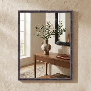 May include: A rectangular wall mirror featuring a dark, textured frame with a bamboo-inspired design. The mirror reflects a wooden console table, a ceramic vase containing olive branches, and a leather-bound book, all set against a wall.