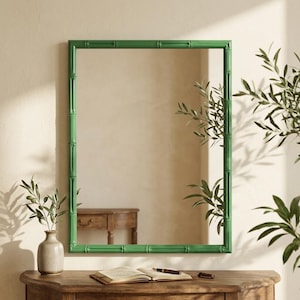May include: A rectangular wall mirror featuring a bright green frame with a textured bamboo-style design. The mirror surface is clear and reflective, showing a reflection of a wooden table and green plant foliage within the room setting.