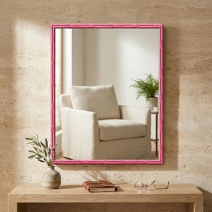 May include: A rectangular wall mirror featuring a bright pink frame with a textured, bamboo-inspired design. The mirror reflects a room interior containing a beige upholstered armchair and a green potted plant on a small side table.