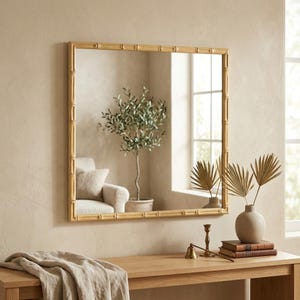 May include: A square wall mirror featuring a light brown frame with a textured bamboo-style design. The mirror surface reflects a room interior with a potted plant, a window, and a chair, providing a clear and reflective glass surface.