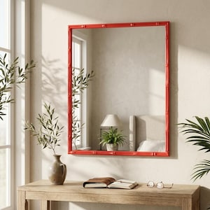 May include: A rectangular wall mirror featuring a bright red frame with a textured bamboo-style design. The mirror reflects a room interior with a lamp and a potted plant, and it is mounted on a plain wall above a wooden console table.