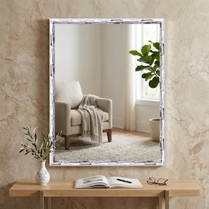 May include: A rectangular wall mirror featuring a silver-toned frame with a textured, bamboo-inspired design. The mirror reflects a room interior containing a beige armchair with a throw blanket and a potted plant near a window.