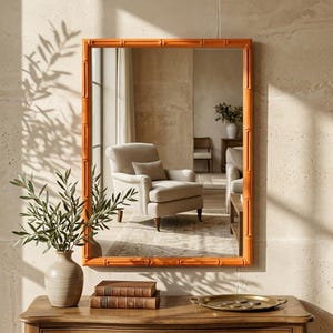 May include: A rectangular wall mirror with a bright orange frame featuring a textured bamboo-style design. The mirror reflects a room with a beige armchair, a wooden side table, and a potted plant. The frame has a smooth, glossy finish.
