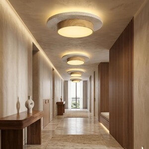May include: A series of circular flush mount ceiling lights installed in a hallway. Each fixture features a wide, textured light grey concrete base, a central light-toned wood ring, and a flat, circular white diffuser emitting a warm glow.