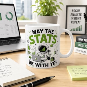May include: A white ceramic mug featuring a graphic of an astronaut working on a laptop surrounded by charts and graphs. The text reads MAY THE STATS BE WITH YOU in green and black lettering. The design includes small planets and a coffee mug.