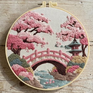 Può includere: Un telaio da ricamo circolare contenente un pannello in tessuto con un paesaggio cucito a mano. Il design presenta un ciliegio in fiore rosa, un ponte ad arco rosso sopra un ruscello blu e una piccola pagoda su uno sfondo chiaro.