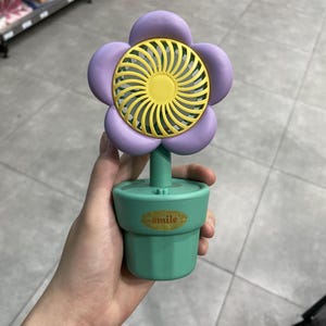 May include: A portable handheld fan shaped like a purple flower with a yellow centre. The fan is mounted on a green base designed to look like a small flower pot. A gold-coloured oval label with the word smile is centred on the front of the base.