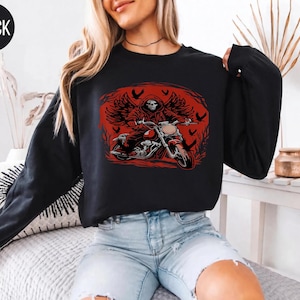 May include: A black long-sleeve sweatshirt featuring a graphic print of a hooded skeleton with wings riding a motorcycle. The design is set against a circular red background with silhouettes of birds. The word BLACK is printed in a circle at the top left.
