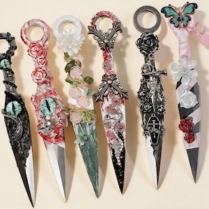 May include: Six decorative kunai knives featuring varied embellishments including a green eye, red roses, white flowers, a skull, a coffin, and a butterfly. Each knife has a ring pommel and a pointed blade, decorated with paint, charms, and fabric.
