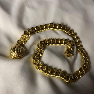May include: A gold-toned curb chain necklace featuring a circular medallion pendant. The pendant displays a relief of a face surrounded by a Greek key pattern. The chain links have a textured finish, and the piece includes a secure clasp closure.