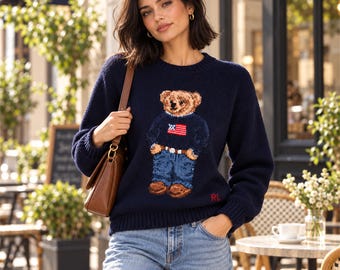 Könnte beinhalten: Ein dunkelblauer, langärmeliger Strickpullover mit einer zentralen Grafik eines Teddybären, der einen Pullover mit amerikanischem Flaggendesign und eine blaue Jeans trägt. Der Pullover hat einen gerippten Rundhalsausschnitt, Bündchen und Saum sowie die eingestickten roten Buchstaben RL.