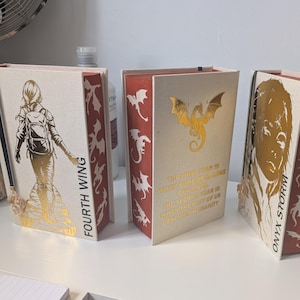 Puede incluir: Tres libros de tapa dura con cubiertas de color crema y bordes de página rojos. El primer libro presenta una figura de lámina dorada y el texto Fourth Wing. El segundo libro muestra un dragón dorado y texto. El tercero muestra un retrato dorado y Onyx Storm.