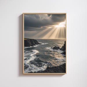 May include: A framed wall art print featuring a dramatic coastal landscape with dark, turbulent ocean waves crashing against jagged black cliffs under a cloudy sky where bright sunbeams break through the clouds to illuminate the water surface.