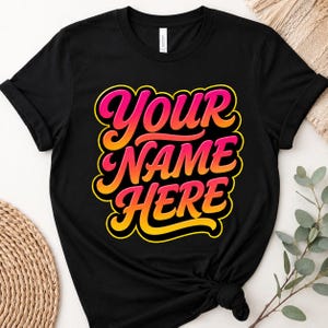 Puede incluir: Una camiseta negra de manga corta con un gráfico central que muestra el texto "YOUR NAME HERE" en una fuente cursiva y audaz de estilo retro. Las letras presentan un degradado vibrante que va del rosa brillante arriba al naranja y amarillo.