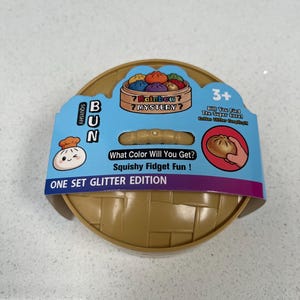 Puede incluir: Un recipiente redondo de plástico color canela con forma de vaporera de bambú, con una funda de cartón azul etiquetada como Squishy Bun Rainbow Mystery. El empaque incluye texto, un dumpling de dibujos animados y un gráfico de una mano.