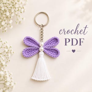 May include: A crochet dragonfly keychain featuring four lavender crocheted wings, two white pearl beads, and a white tassel. The keychain includes a silver metal ring and chain. The text "crochet PDF" is visible on the right side of the object.