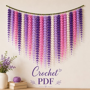 May include: A wall hanging featuring multiple vertical strands of crocheted flowers in shades of purple, pink, and white, suspended from a green cord. The text "Crochet PDF" is printed below the hanging, accompanied by small decorative leaf icons.