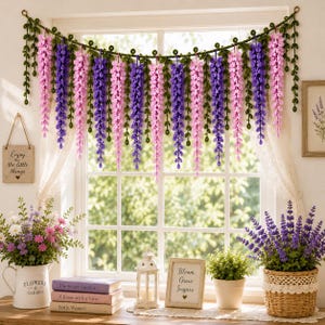 May include: A handmade crochet wisteria garland featuring alternating long, hanging clusters of purple and pink flowers attached to a green vine with small leaves. The decorative floral piece is draped across the top of a window frame.