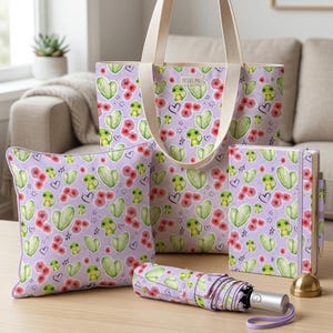 Può includere: Una collezione di articoli con un motivo ripetuto di cactus verdi, fiori rossi e rane dei cartoni animati su sfondo lavanda. Gli articoli includono una borsa tote, un cuscino, un diario, una penna e un ombrello. La borsa porta il testo "Desert Pal".