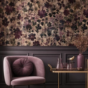May include: A mauve velvet armchair with a round pleated cushion, positioned next to a gold metal bar cart holding a ribbed mauve vase, a small gold bird figurine, and several glass vessels against a dark wall with floral wallpaper.