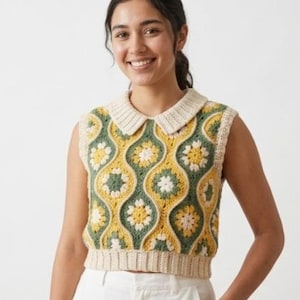 May include: A sleeveless crochet vest featuring a repeating pattern of yellow and white floral motifs set against a sage green background. The garment includes a cream-colored ribbed collar and a matching ribbed hem at the waistline.