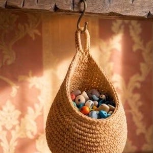 May include: A beige, teardrop-shaped crochet hanging basket suspended from a metal S-hook. The basket is constructed with a dense, textured stitch pattern and features a single top loop for hanging. It is filled with a collection of small, multi-colored beads.