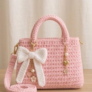 May include: A light pink crocheted handbag featuring a structured rectangular body and two top handles. A large white crocheted bow is attached to the front, accented with a gold-toned charm chain featuring a heart, a pearl, and a flower pendant.