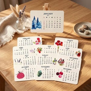 May include: A 2027 desk calendar set featuring twelve individual rectangular cards, each displaying a month with watercolor illustrations. One card rests on a small wooden easel, while the others are arranged below. The designs include pine trees, flowers, lavender, and seasonal motifs printed on off-white cardstock.