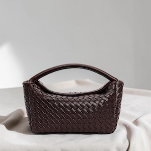 Puede incluir: Un bolso de mano de color marrón oscuro con un exterior de cuero trenzado que presenta un patrón de rejilla distintivo. El bolso tiene una silueta superior curva y un asa única integrada que se arquea sobre el centro, ofreciendo un aspecto estructurado y compacto.