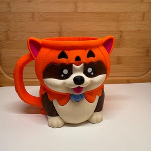 Könnte beinhalten: Ein Becher in Form eines Waschbären, der ein orangefarbenes Kürbiskostüm trägt. Der Waschbär hat braunes und cremefarbenes Fell, große schwarze Augen und eine kleine rosa Zunge. Die Kürbiskapuze hat schwarze dreieckige Augen und einen orangefarbenen Henkel.