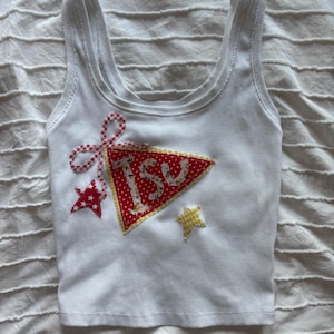 May include: A white ribbed tank top featuring a red pennant-shaped patch with white polka dots and the letters "tsu" in a floral print. The design includes a red and white gingham bow, a red star with white stars, and a yellow checkered star.