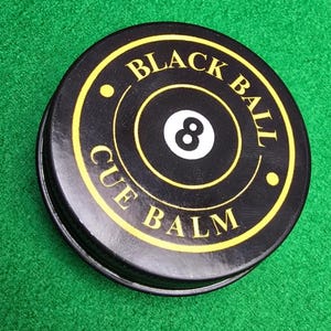 May include: A black cue balm tin with yellow text and a white number 8. The text on the tin reads "BLACK BALL CUE BALM". The round tin is on a green felt surface, often found on a snooker or pool table.