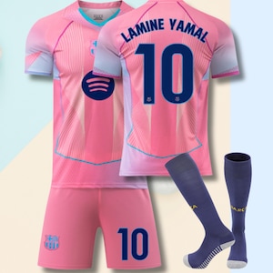 May include: A pink and light blue football kit featuring the name LAMINE YAMAL and the number 10 in dark blue. The shirt includes a Spotify logo, a Barcelona crest, and matching pink shorts with the number 10, plus dark blue football socks.