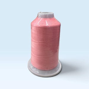 May include: A large spool of soft pink sewing thread wrapped in clear plastic film. The thread is wound tightly around a white cylindrical plastic core with a wide base, creating a uniform and smooth texture across the entire surface of the spool.