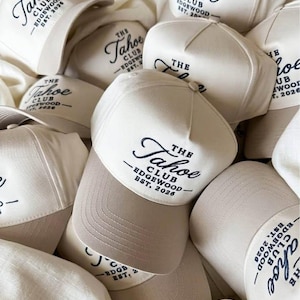 May include: A collection of cream-coloured baseball caps featuring the embroidered text "THE Tahoe CLUB EDGEWOOD EST. 2026" in dark navy blue. Each cap has a structured front panel and a curved brim, stacked together in a pile.
