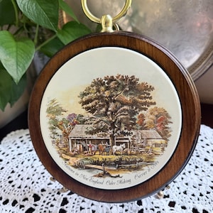 May include: A round ceramic wall plaque featuring a Currier & Ives print titled Autumn in New England Cider Making. The artwork depicts a rural scene with a large tree, a building, and oxen. It is set in a dark wood frame with a gold metal ring.