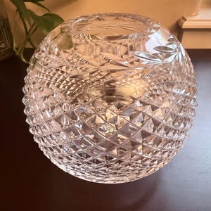 May include: A clear, spherical crystal bowl featuring an intricate, multi-faceted diamond cut pattern across its entire exterior surface. The glass vessel has a wide, circular opening at the top and a smooth, polished rim, reflecting light through its geometric design.