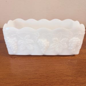 May include: A rectangular white milk glass planter featuring a scalloped top edge and an embossed pattern of grape clusters and vine leaves wrapping around the exterior sides. The vessel has a smooth, opaque finish and a consistent white color.