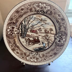 May include: A round ceramic plate featuring a brown and white transferware pattern. The center depicts a winter landscape with a horse-drawn sleigh, trees, and a house. The wide rim is decorated with a detailed floral and lattice border design.
