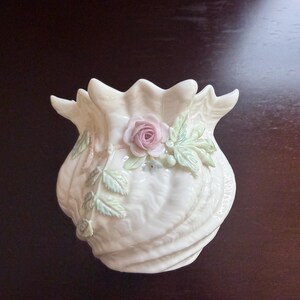 May include: A cream-colored porcelain vase with a scalloped, shell-like rim and a textured, swirling body. The vase is decorated with a three-dimensional, sculpted pale pink rose blossom and delicate green leaves attached to the exterior surface.