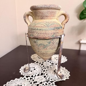 May include: A beige and green ceramic amphora vase with two handles, featuring embossed scroll patterns and a speckled texture. The vase is held in a dark metal stand with three legs, designed to support its pointed, conical base.