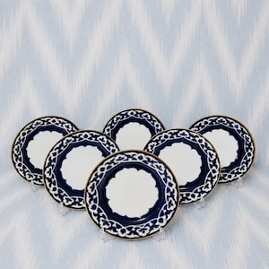 May include: A set of five round ceramic plates featuring a solid white center surrounded by a deep navy blue border with a white floral vine pattern and a thin gold rim. The plates are arranged in a staggered formation on a light blue background.