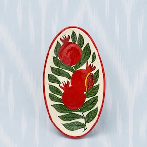 May include: An oval ceramic dish with a cream-colored base and a red rim. The surface features a hand-painted design of three bright red pomegranates with green leaves and stems arranged vertically along the center of the dish.