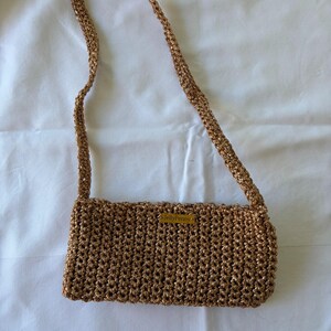 May include: A rectangular shoulder bag crafted from metallic bronze-colored yarn in a textured crochet stitch. The bag features a single long strap and a small rectangular label on the front with the text JellyPenny printed in black letters.